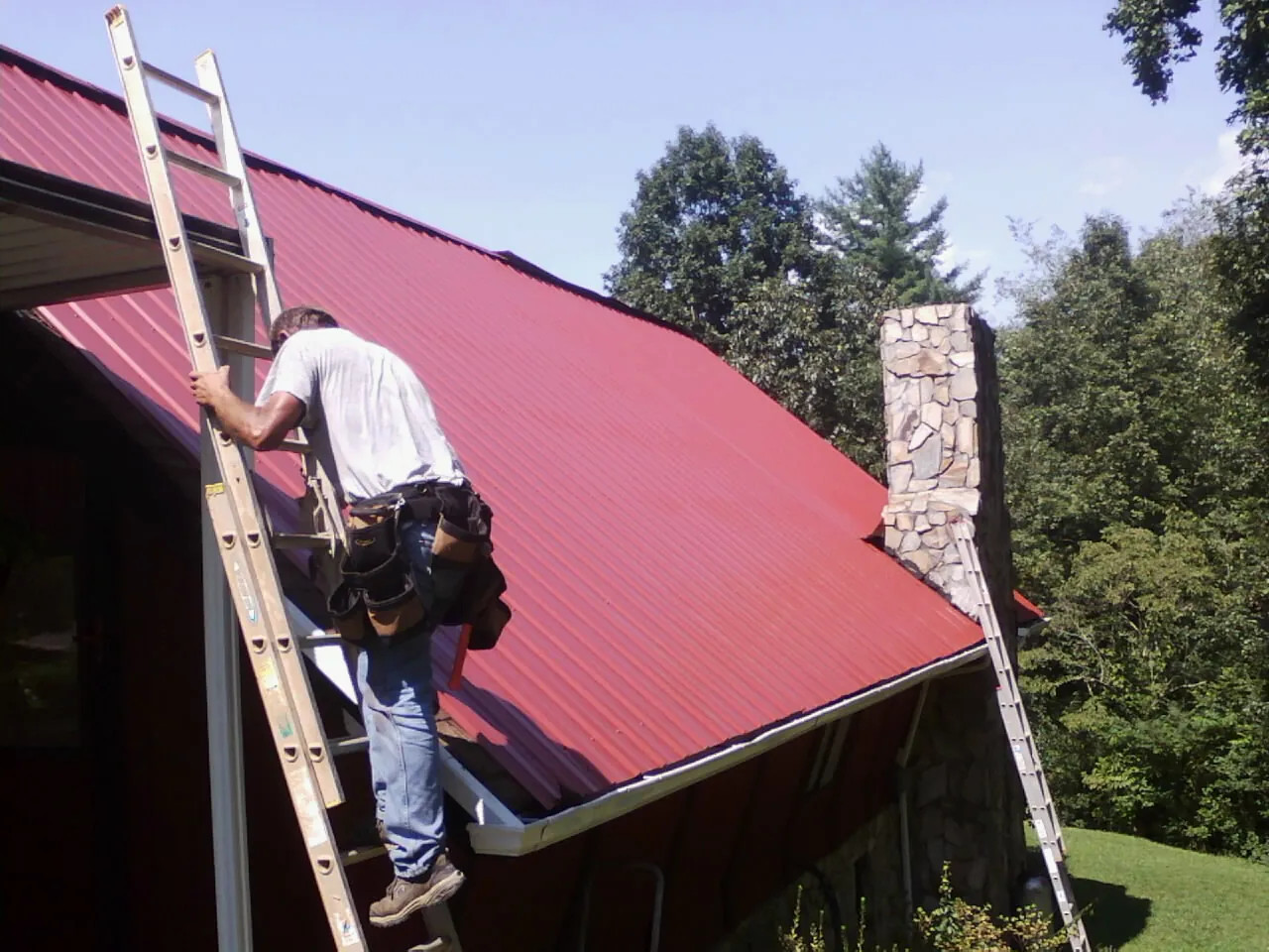Roofer on a ladder beside a red metal roof and stone chimney — Soffit & Fascia Repair by Roofing Brothers in North Versailles